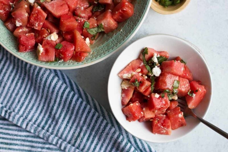 Top view of an individual serving of watermelon basil salad alongside a larger serving bowl of more salad.