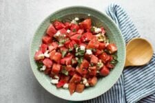 A wooden spoon rests on a striped linen napkin beside a decorative bowl filled with watermelon basil salad.