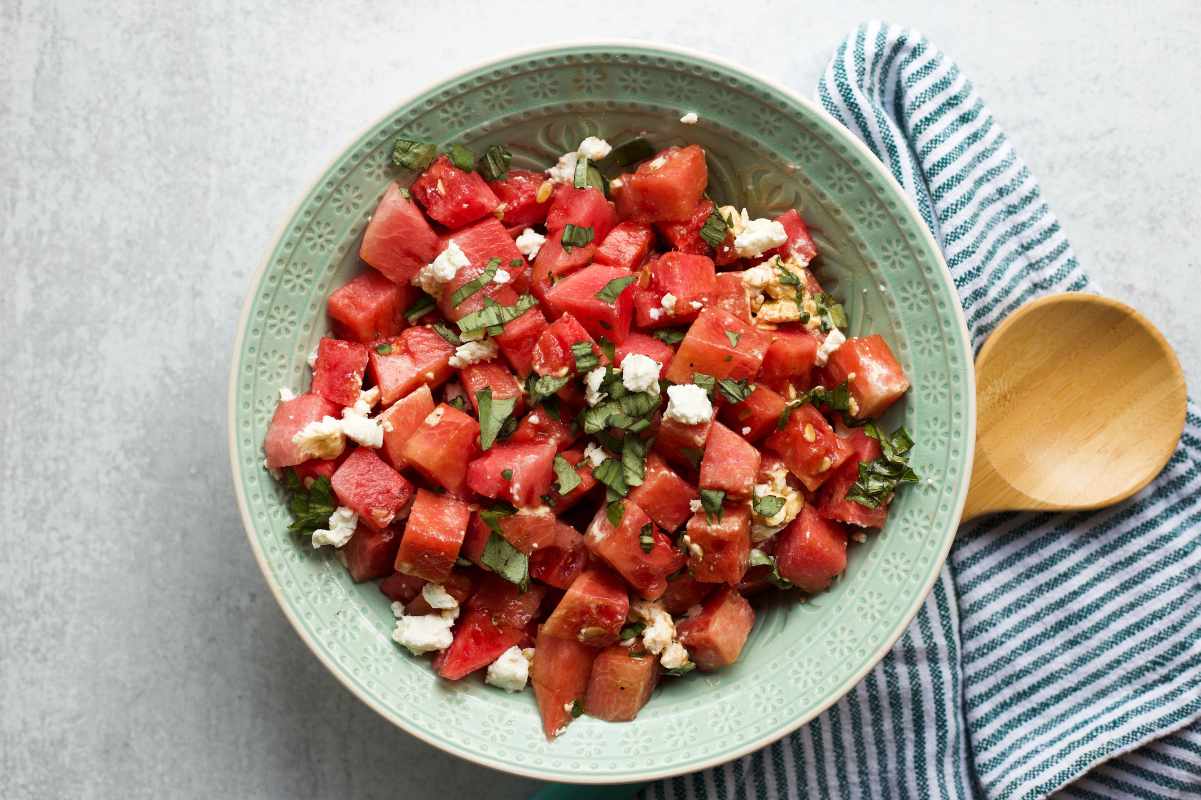 A decorative bowl filled with watermelon basil salad with a striped linen napkin beside it.