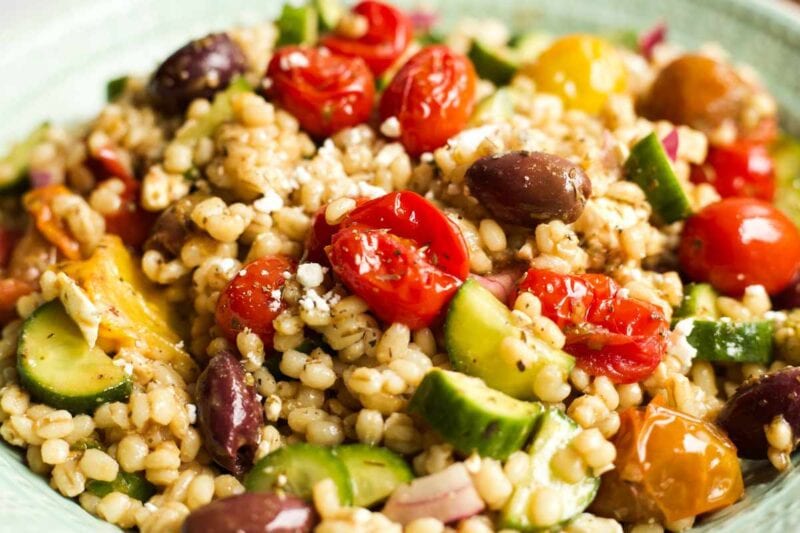 Close view of greek barley salad with pan roasted tomatoes, crumbled feta, sliced cucumbers, and kalamata olives.