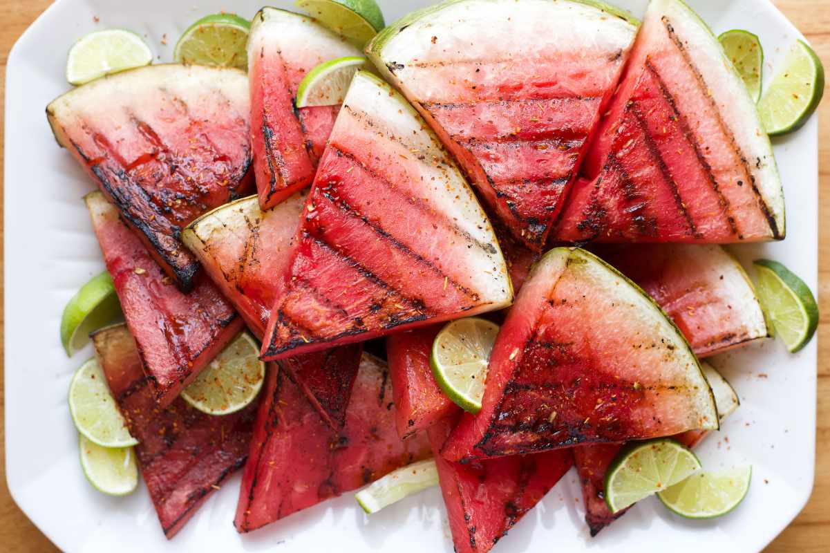 Lime slices are nestled among grilled watermelon slices sprinkled with tajin on a white dish.