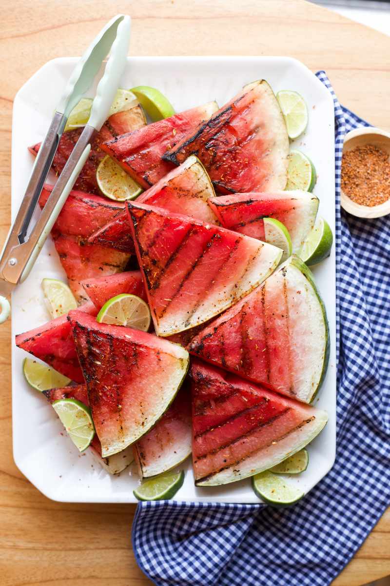 Overhead of a rectangular white plate piled high with grilled watermelon slices garnished with lime wedges and tajin seasoning.