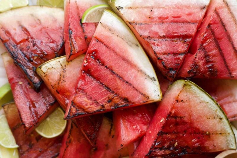 Tight view of grilled watermelon slices piled atop one another with slices of lime nestled among them.