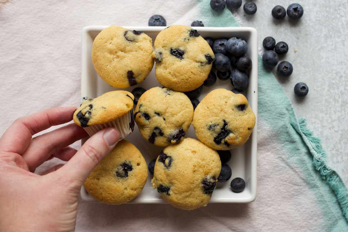 A hand takes a mini muffin from a square serving platter with fresh berries.