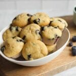 A serving bowl of mini blueberry muffins rests on a serving board with a small bowl of fresh berries beside it.