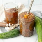 Two home canned jars of sweet relish stand on a counter with fresh pickles around them.