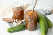 Two home canned jars of sweet relish stand on a counter with fresh pickles around them.