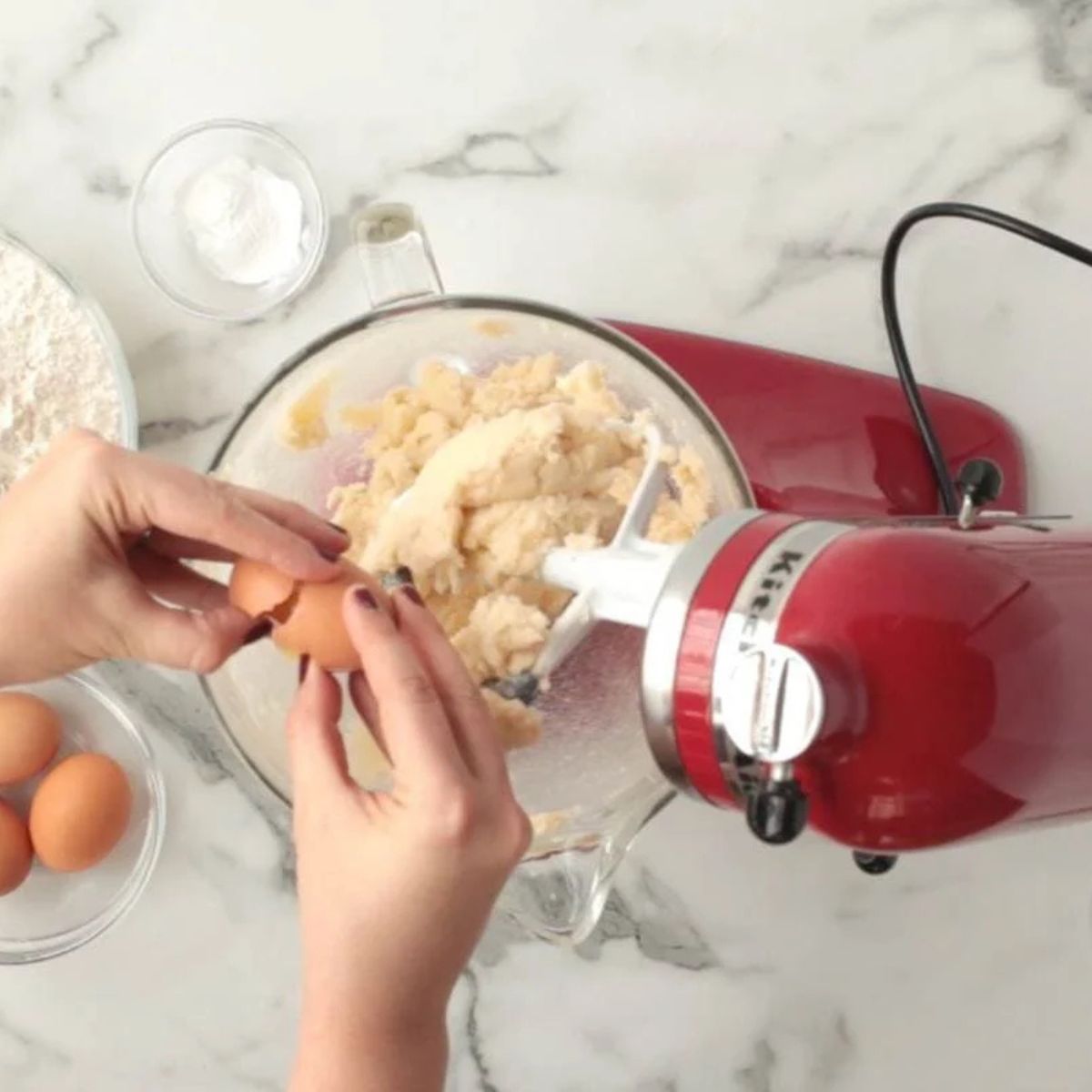 Adding eggs to sugar cookie dough in a red stand mixer on a white marble countertop