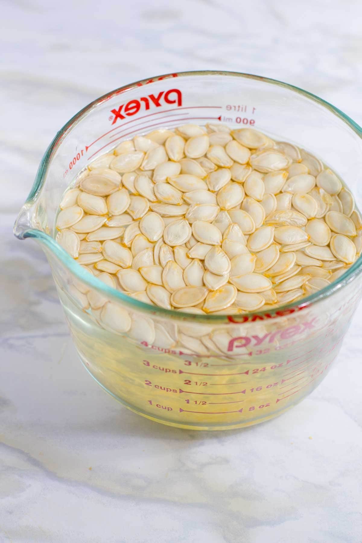 Clean pumpkin seeds float in water inside a clear glass measuring cup on a white countertop
