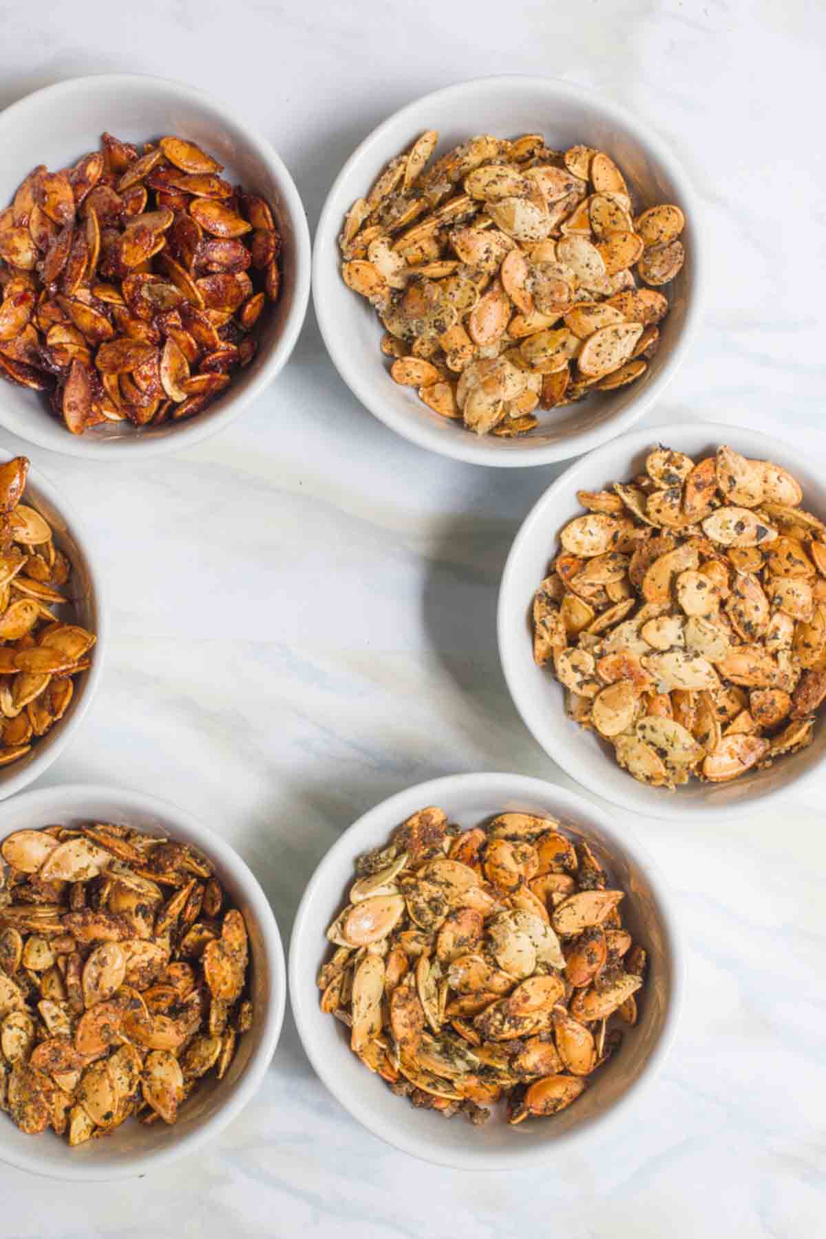 White bowls of seasoned homemade roasted pumpkin seeds sit on a white counter.