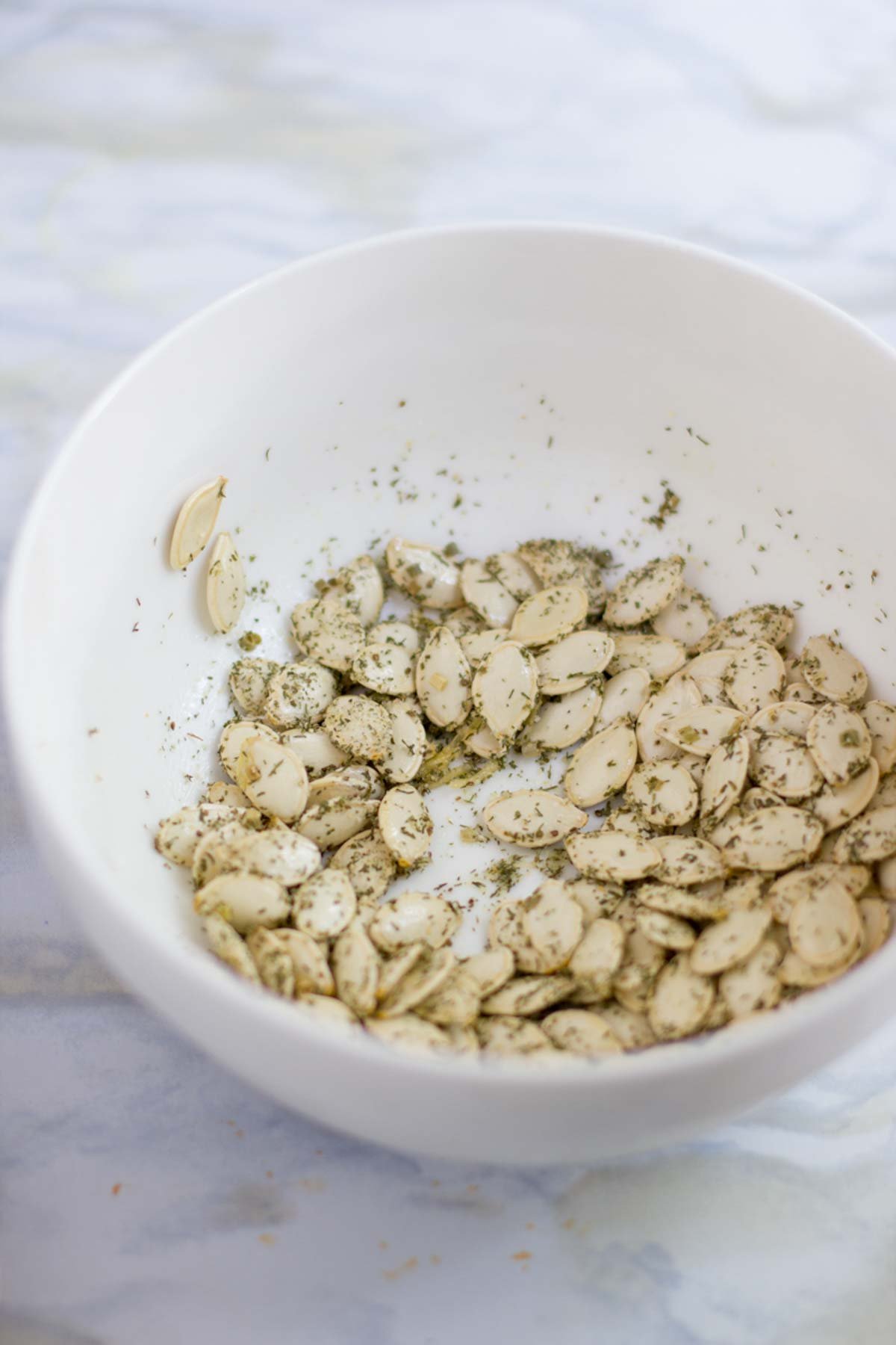 Pumpkin seeds mixed with herbs and oil in a white bowl on a white marble countertop