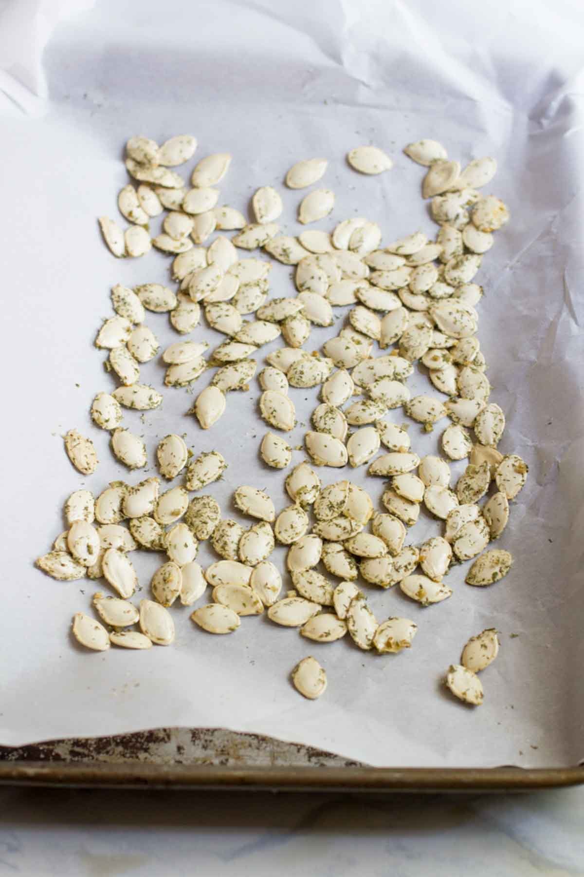 Unroasted pumpkin seeds covered in seasoning spread in one even layer on a baking sheet covered with parchment paper.