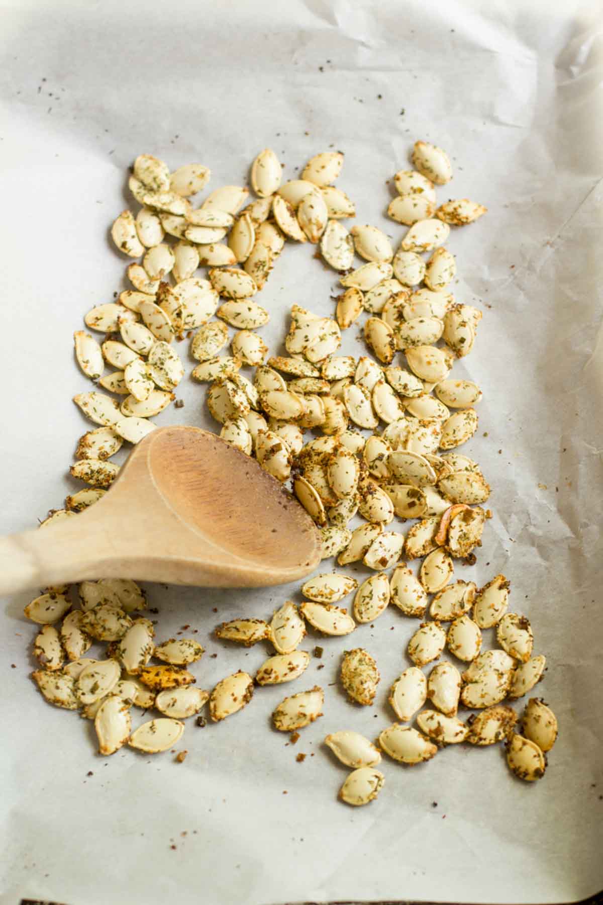 Wooden spoon stirs roasted pumpkin seeds on a parchment lined baking sheet.