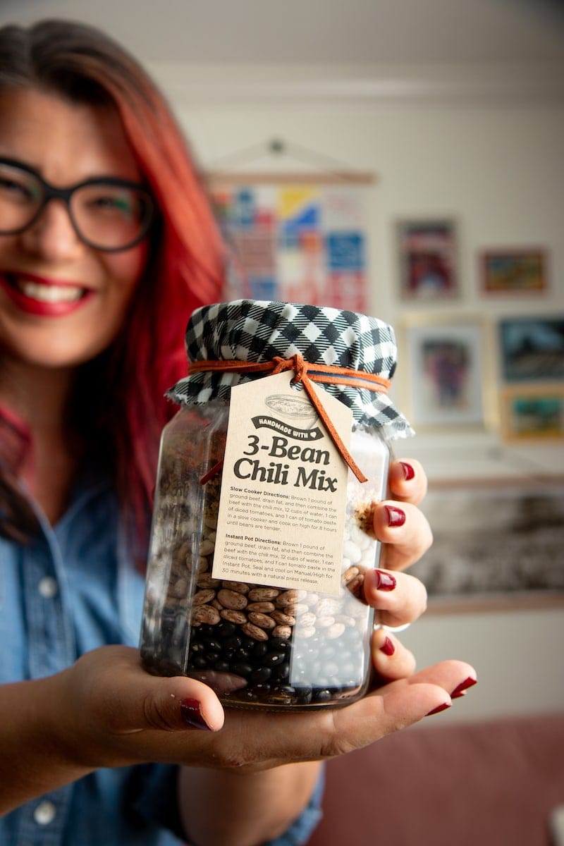 A woman with bright red hair and black glasses smiles in the background blurred while the focus of the image is on a jar she is holding that has 3-Bean Chili Mix inside of it.