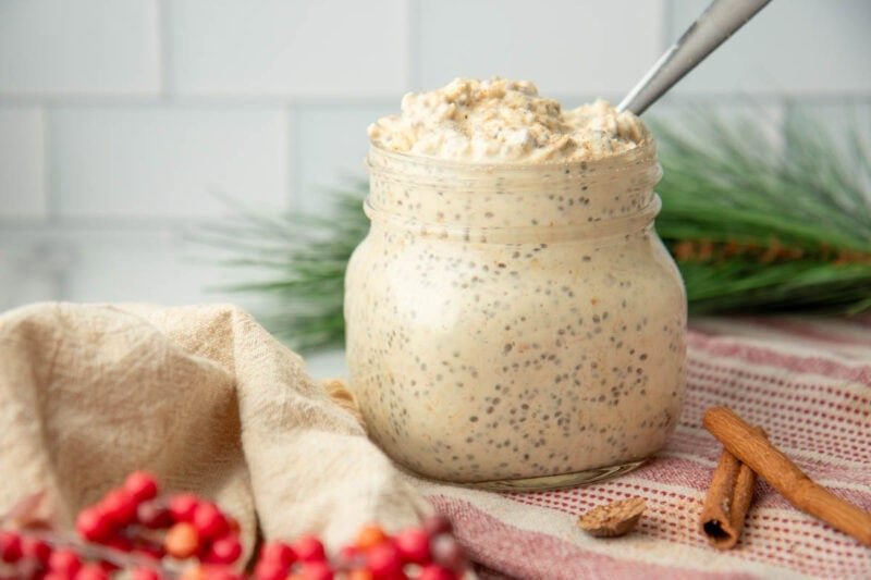 Close view of a jar of creamy holiday oats with red holly berries in the foreground and an evergreen branch in the background.