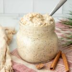 A jar of eggnog overnight oats rests on a counter with two cinnamon sticks and a piece of nutmeg beside it.
