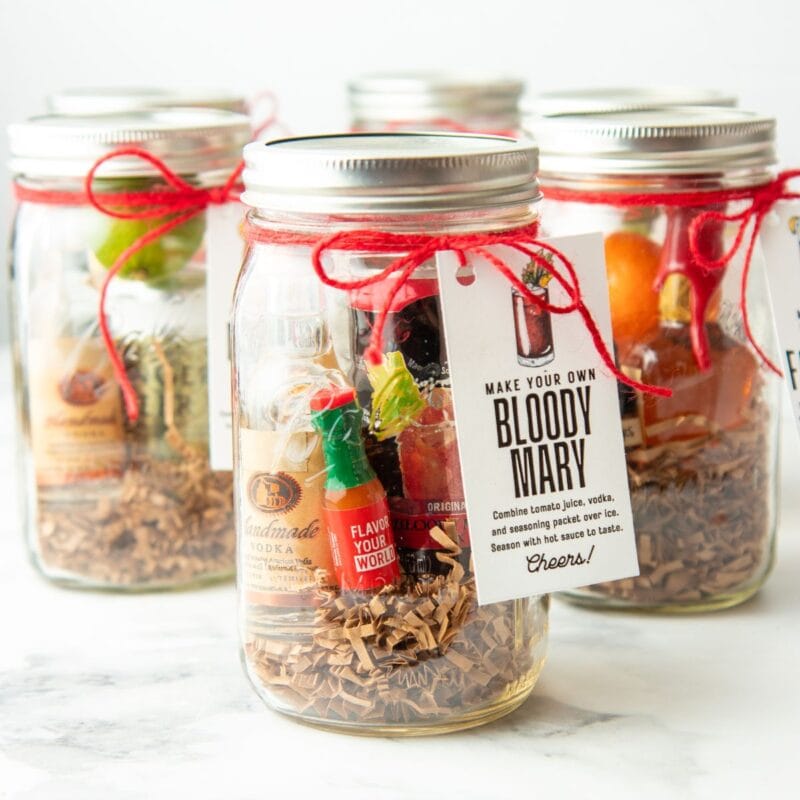 A lineup of clear glass canning jars sit on a white marble countertop with ingredients to make cocktails inside