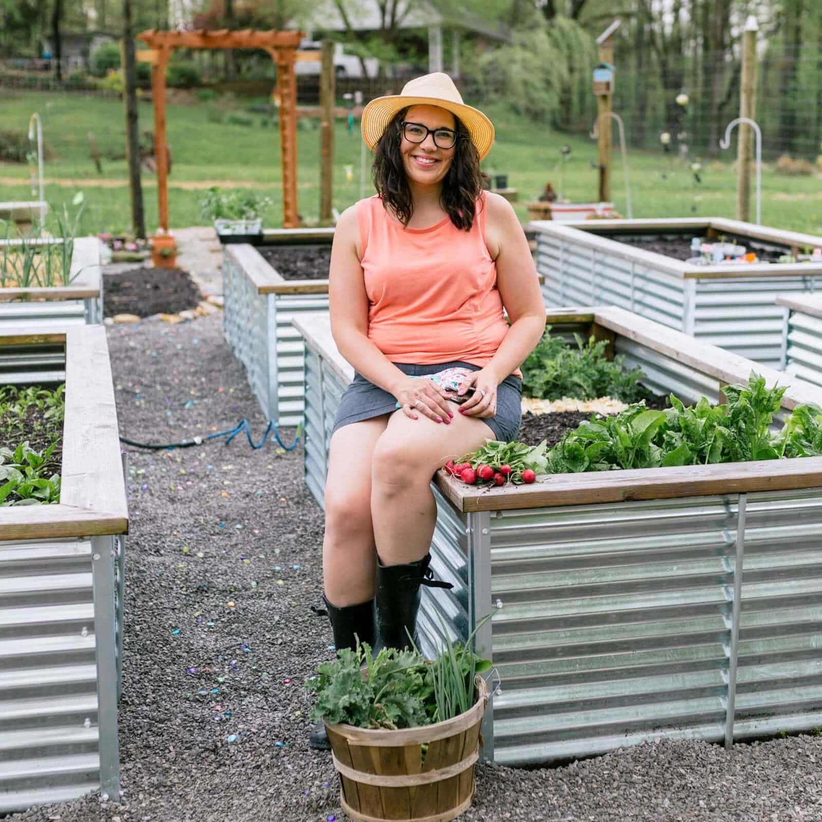 Brunette woman in a coral shirt and a sun hat sitting on the edge of a raised garden bed