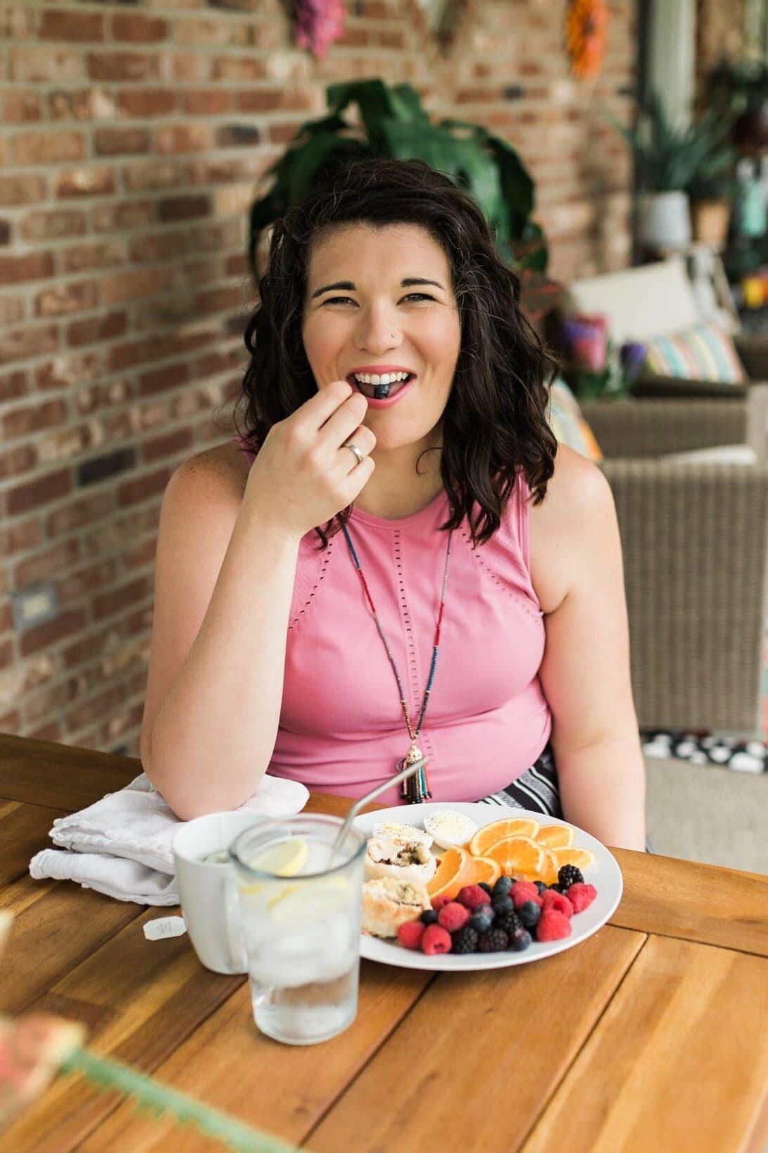 Cassie Johnston sits at a table wearing a pink shirt. She has a plate of food in front of her and smiles while she eats a blueberry.