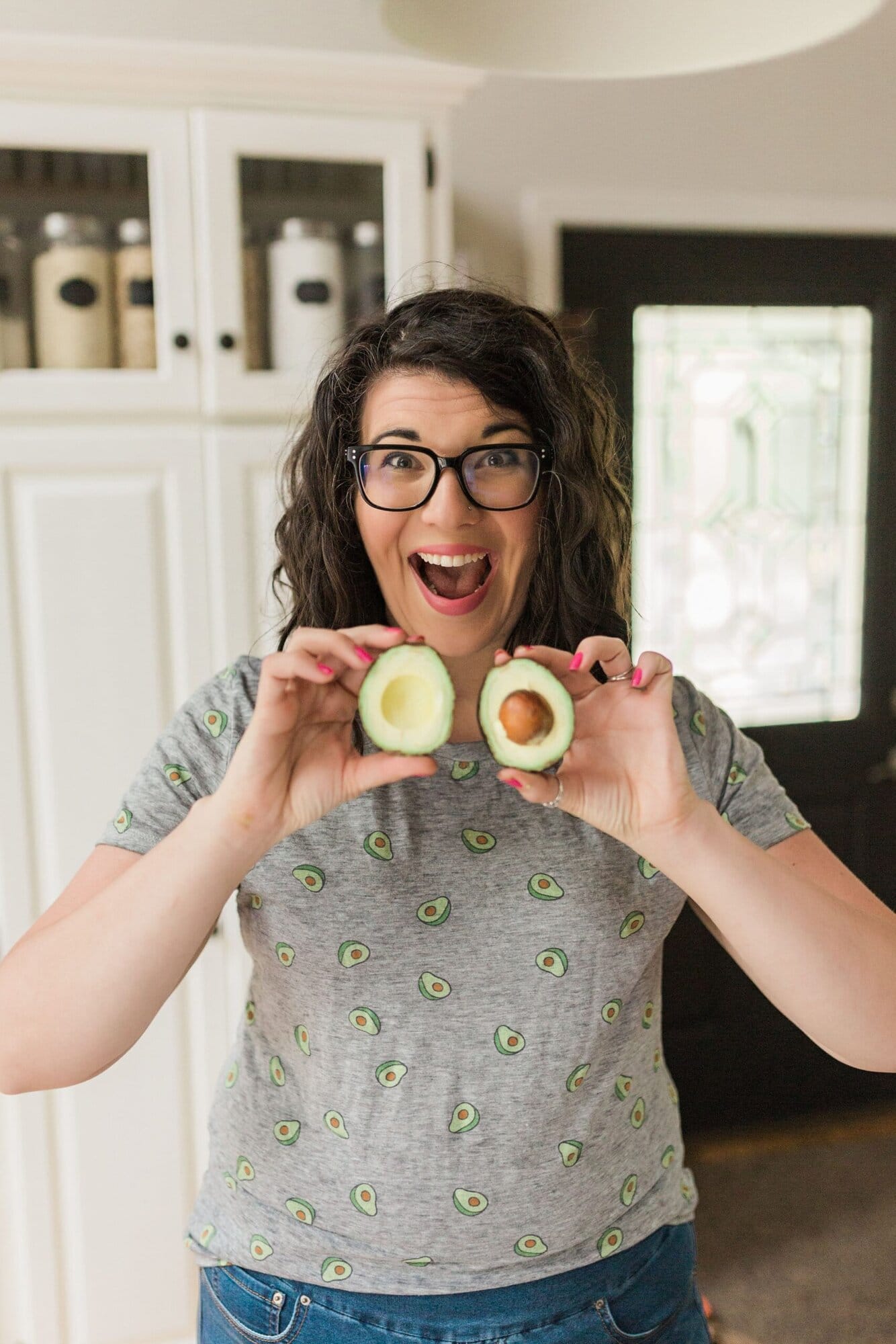Cassie Johnston stands in her white kitchen smiling as she holds a cut avocado. She's wearing a gray avocado shirt and jeans.