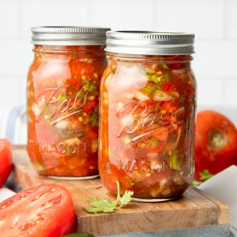 Two mason jars filled with canning salsa on a wooden cutting board surrounded by fresh ingredients.