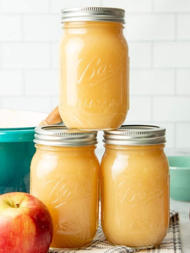 Three canning jars of applesauce are stacked in front of a white kitchen. A red apple sits next to the stack.