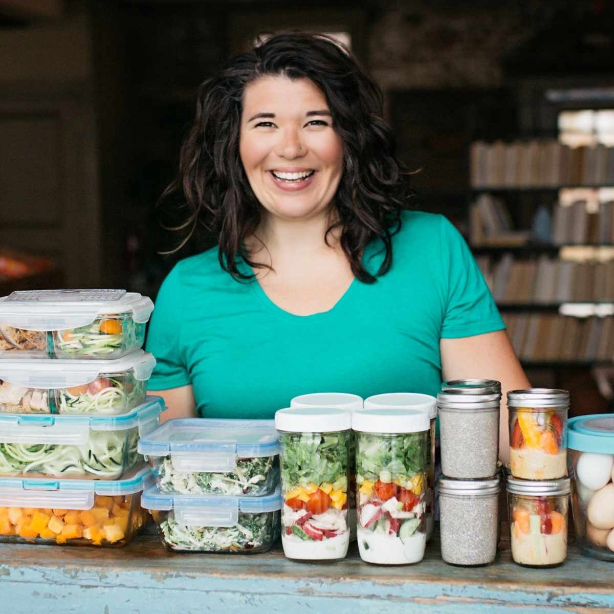 Cassie Johnston smiles in a teal shirt while standing at a table with meal prep containers stacked high in front of her.