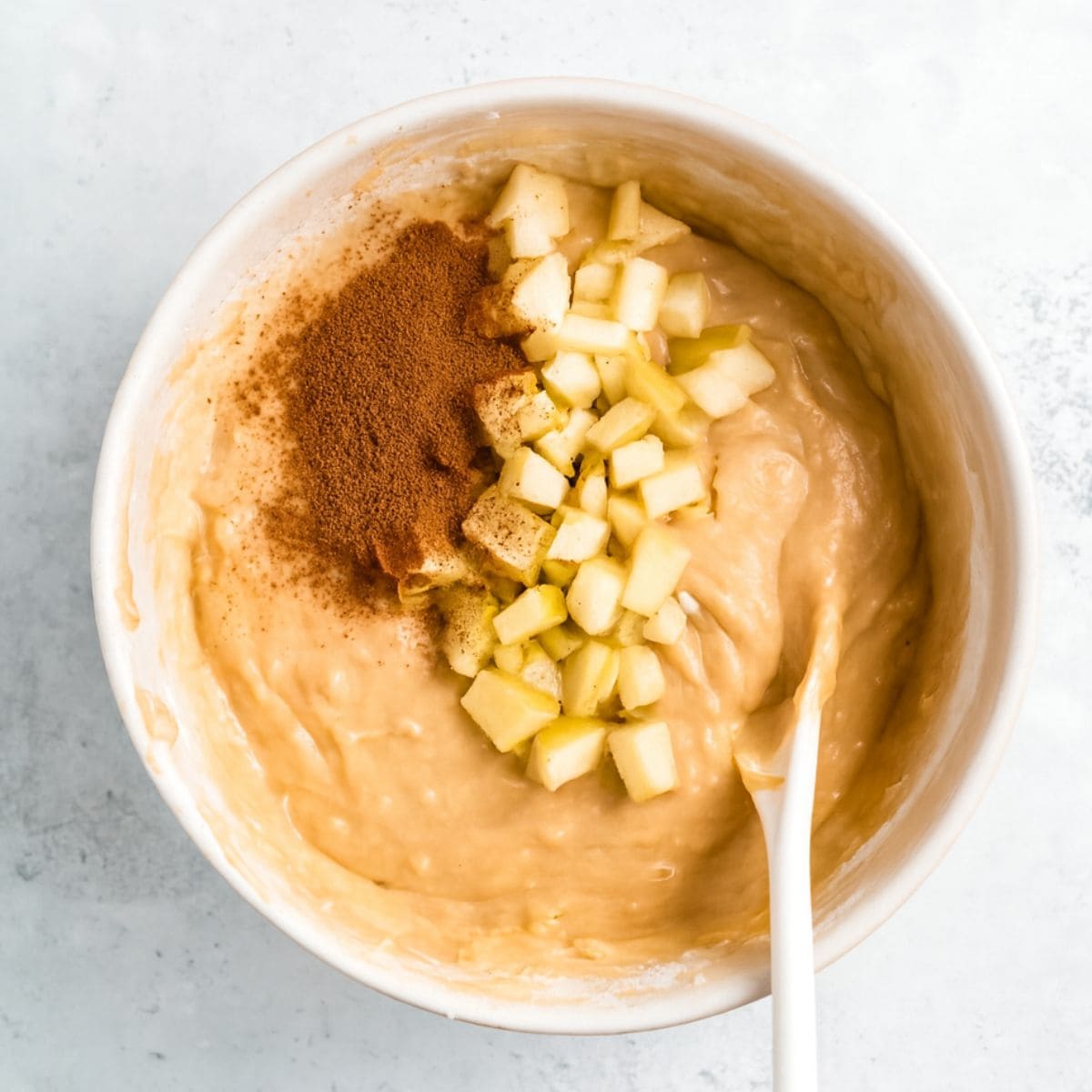 Apples and cinnamon on top of muffin batter in a white bowl with a white spoon.