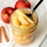A black spoon dips into a jar of canned apple pie filling. Two apples and cinnamon sticks sit nearby.