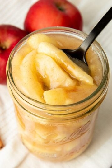 A black spoon dips into a jar of canned apple pie filling. Two apples and cinnamon sticks sit nearby.