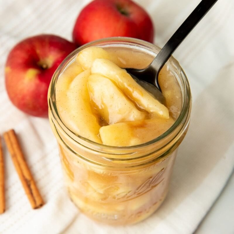A black spoon dips into a jar of canned apple pie filling. Two apples and cinnamon sticks sit nearby.
