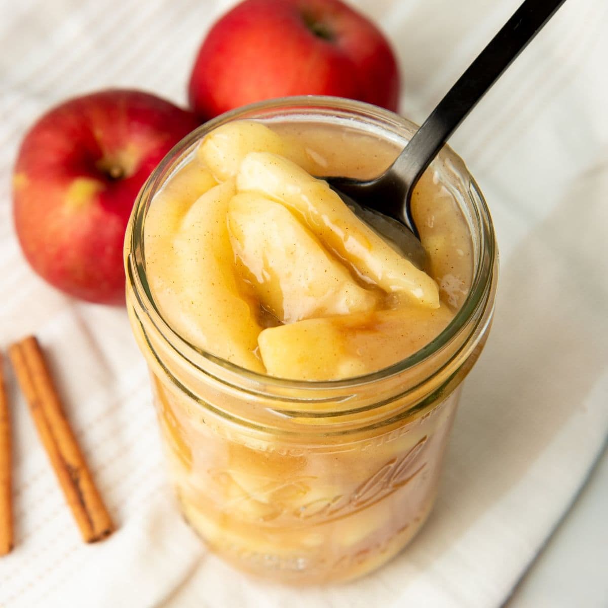 A black spoon dips into a jar of canned apple pie filling. Two apples and cinnamon sticks sit nearby.