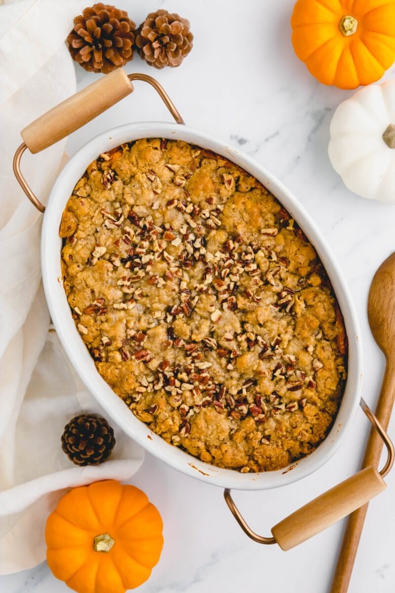 Baked sweet potato casserole in a white pan, surrounded by mini pumpkins and pine cones.