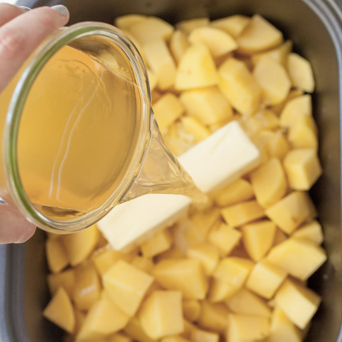 Chicken broth pours over chopped potatoes and butter in the basin of a slow cooker.