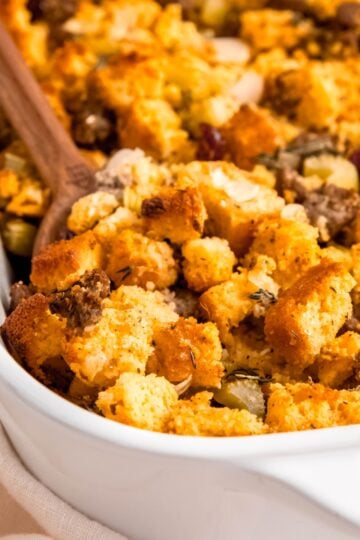 A wooden spoon dips into a casserole dish of cornbread and sausage dressing.