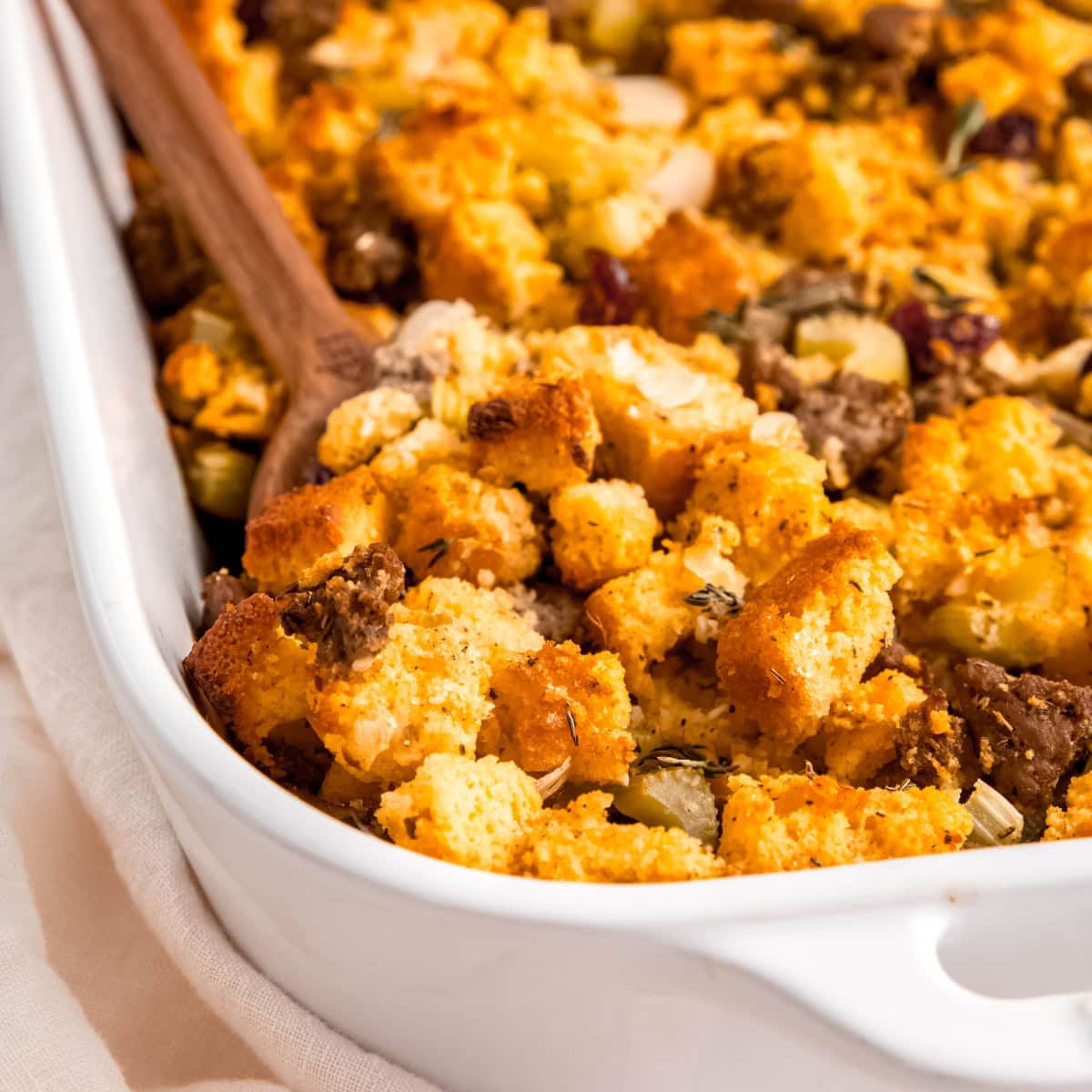 A wooden spoon dips into a casserole dish of cornbread and sausage dressing.