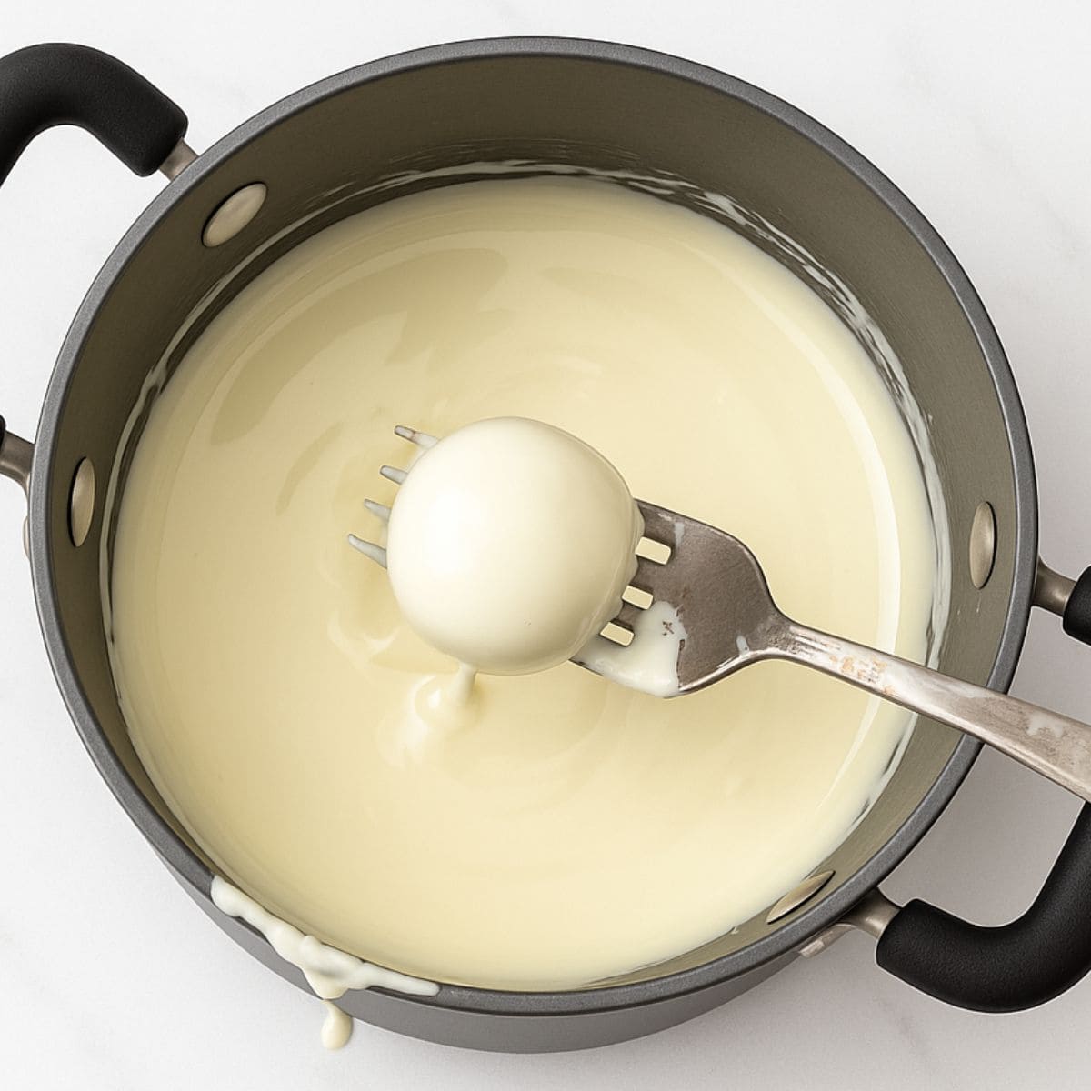 A fork holds a cake pop over a pan of melted white candy coating