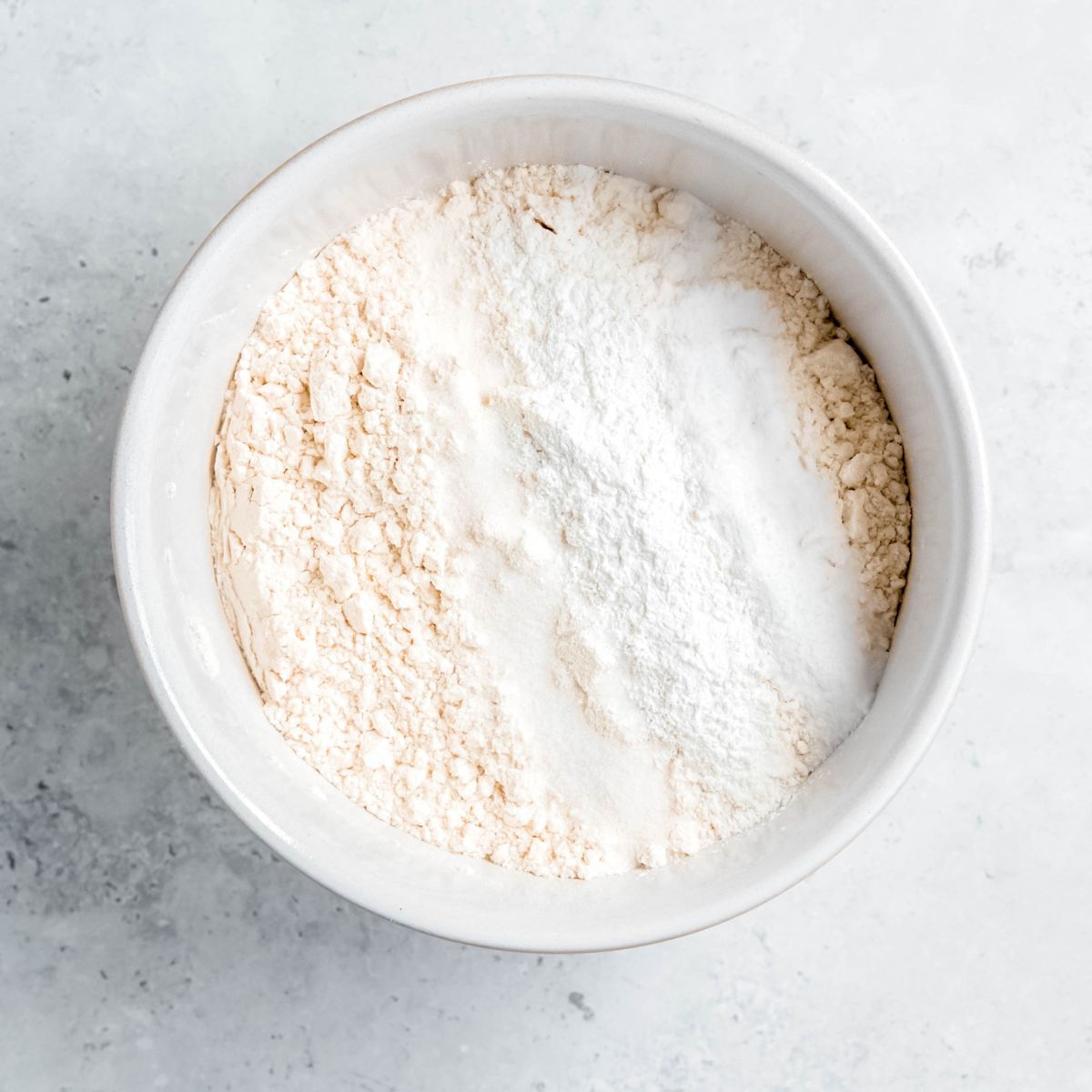 Dry ingredients for muffins in a white mixing bowl on a marbled counter.