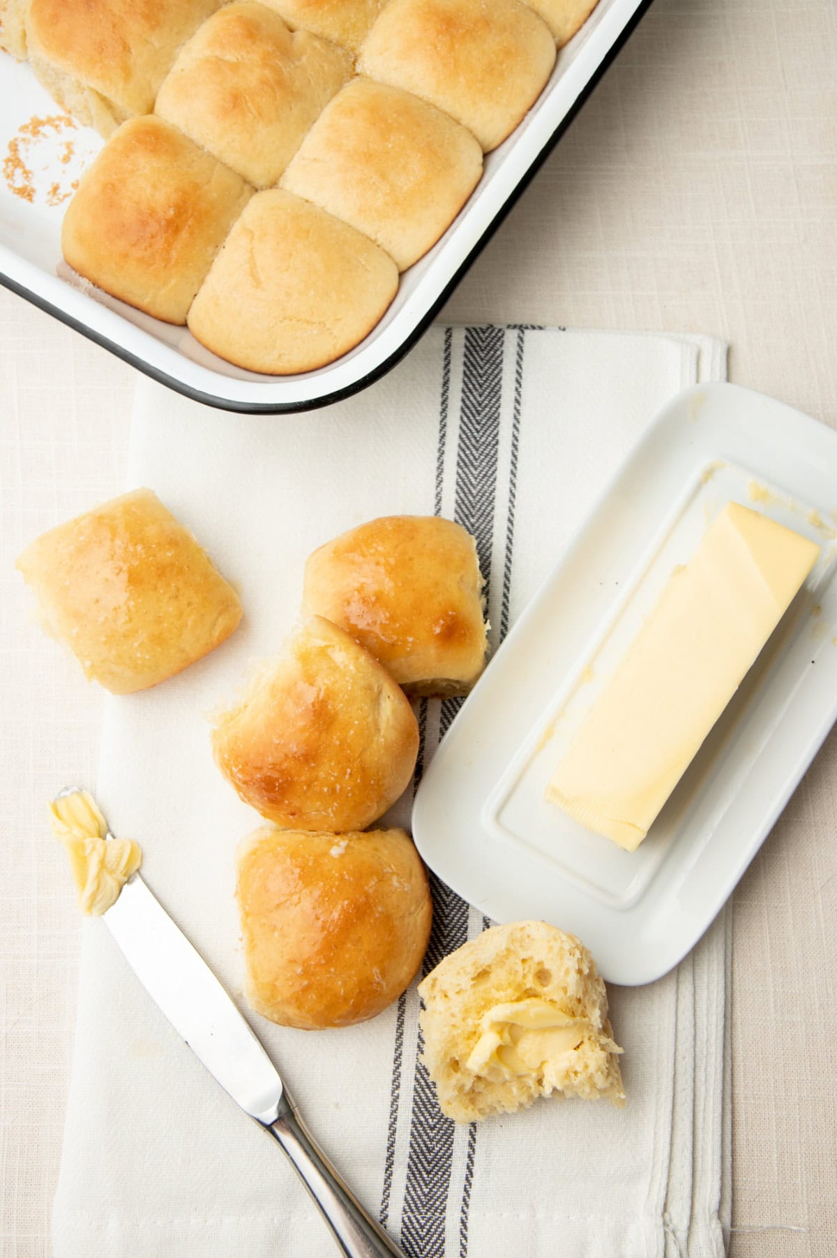 A pan of yeast rolls with some rolls removed to a tea towel. There is a knife and butter dish to spread butter on the rolls.