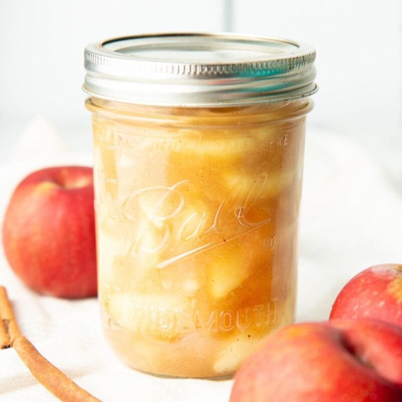 Canned apple pie filling in a sealed jar, surrounded by whole apples.
