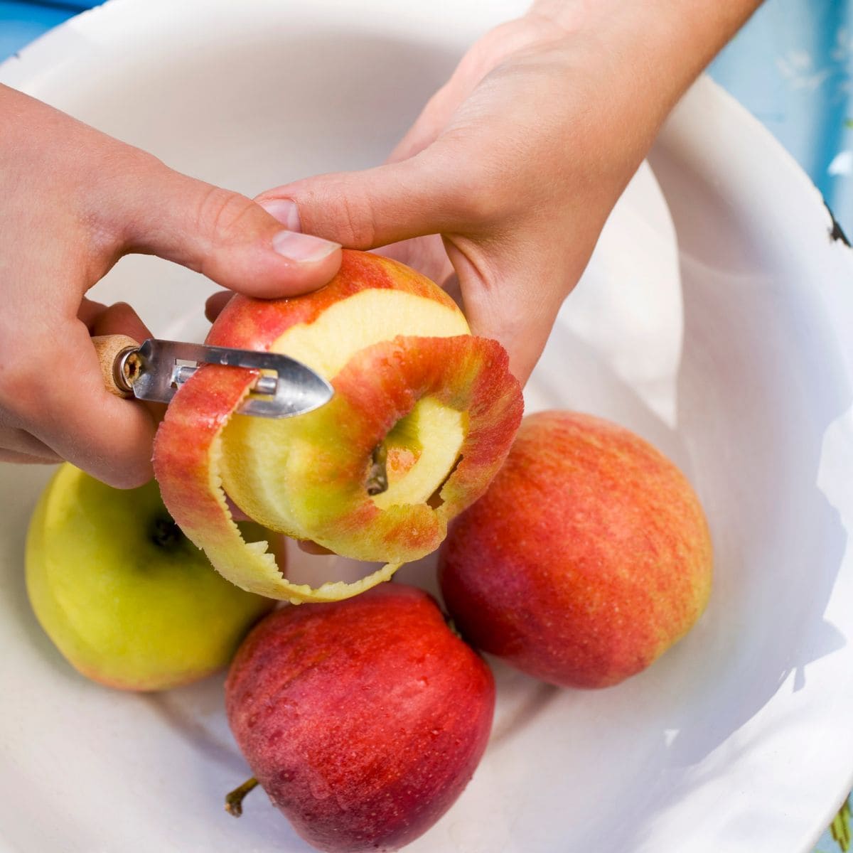 Hands peel a red apple over a white bowl.