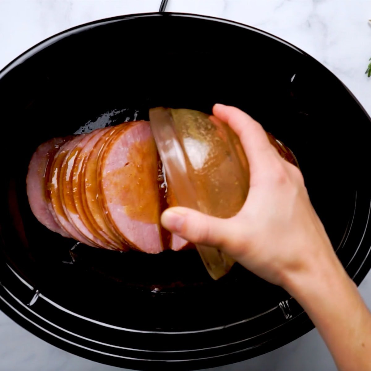 A hand pours honey mustard sauce over a ham in the basin of a slow cooker.