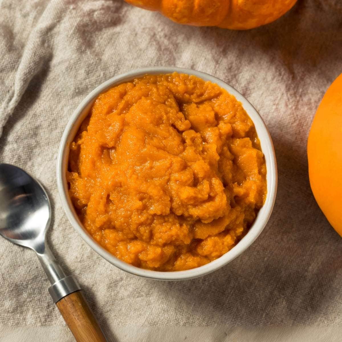 Bowl of pumpkin puree next to a spoon and two pumpkins.