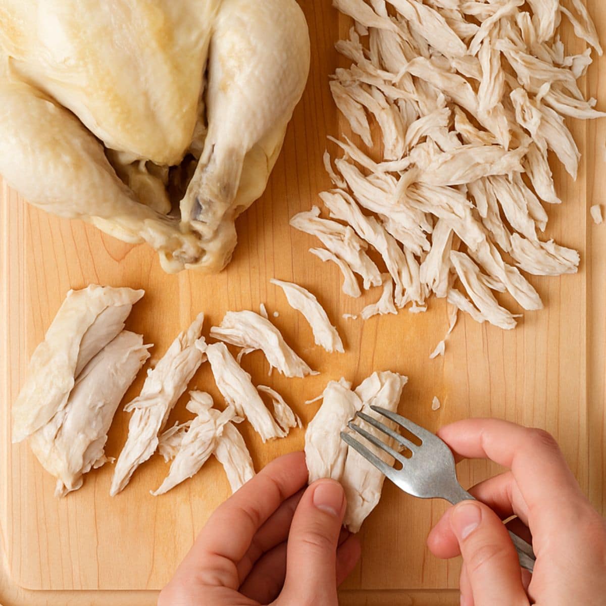 A fork shreds chicken on a wooden cutting board.