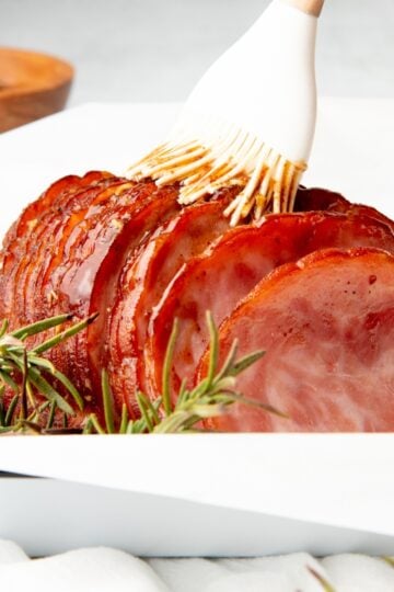A pastry brush paints honey-mustard glaze on a sliced cooked ham.