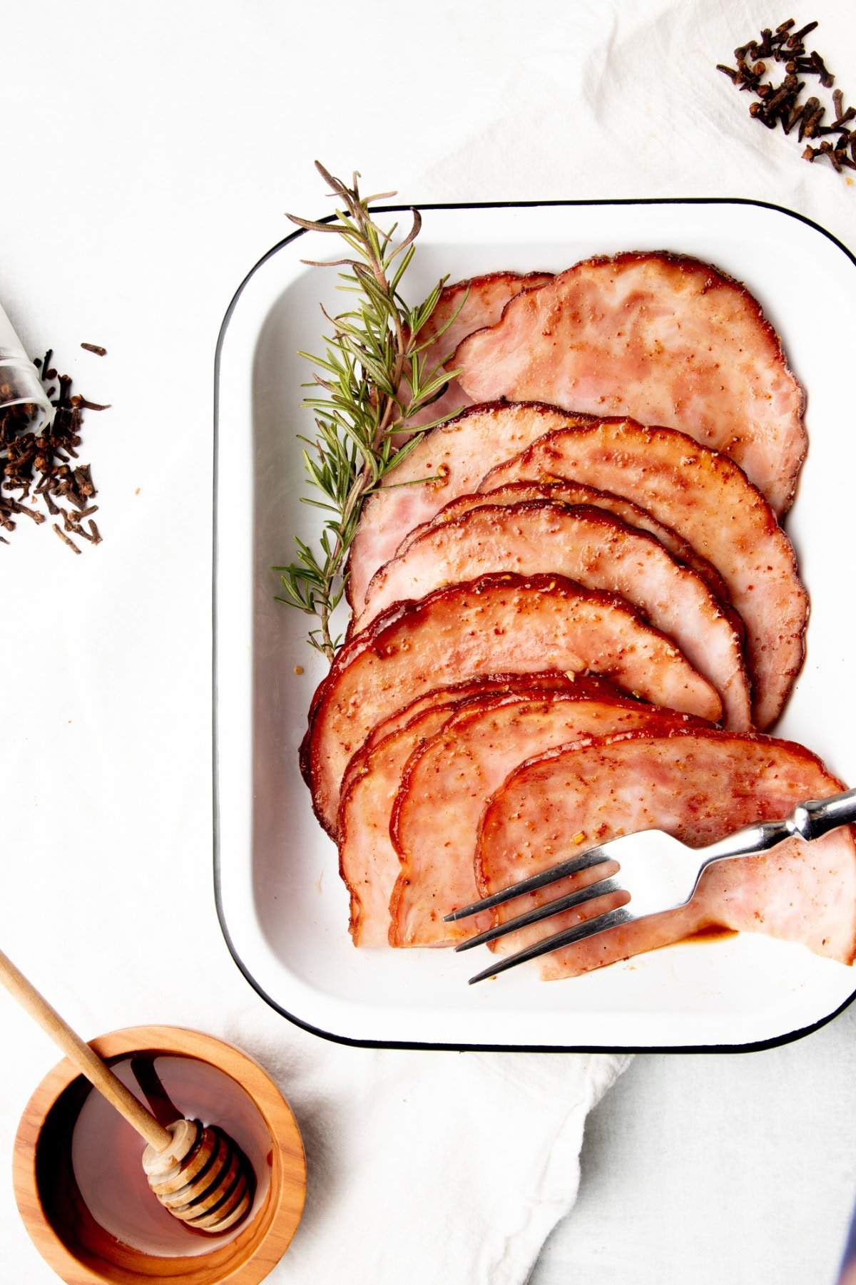 Slices of glazed ham on a white enamel platter with a serving fork.