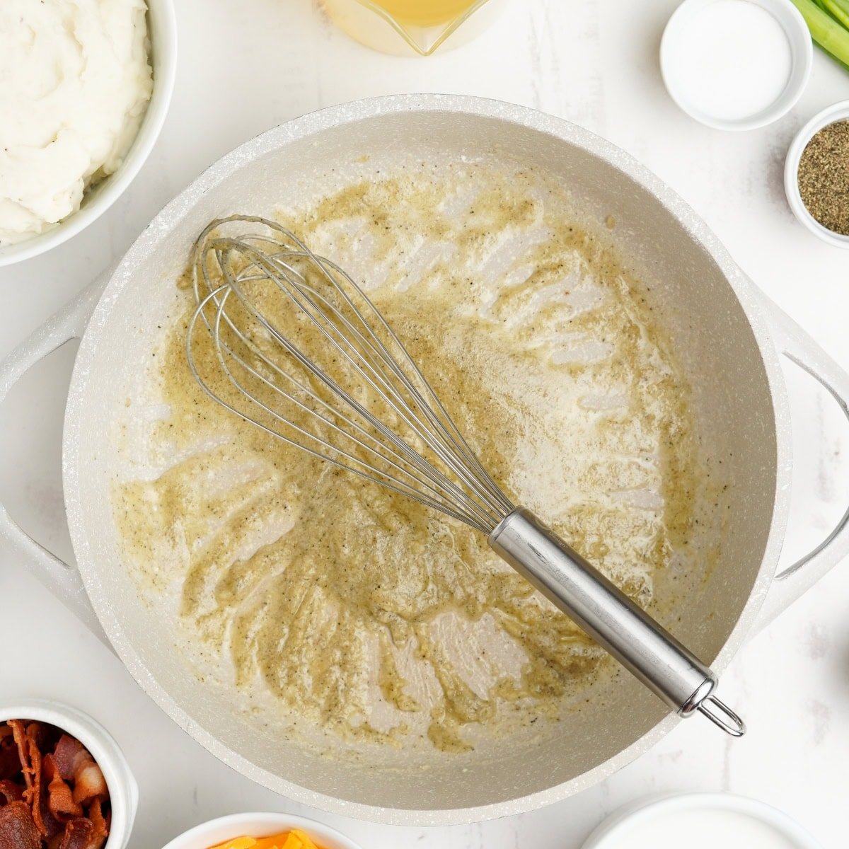 A whisk rests on a roux in a grey pot.