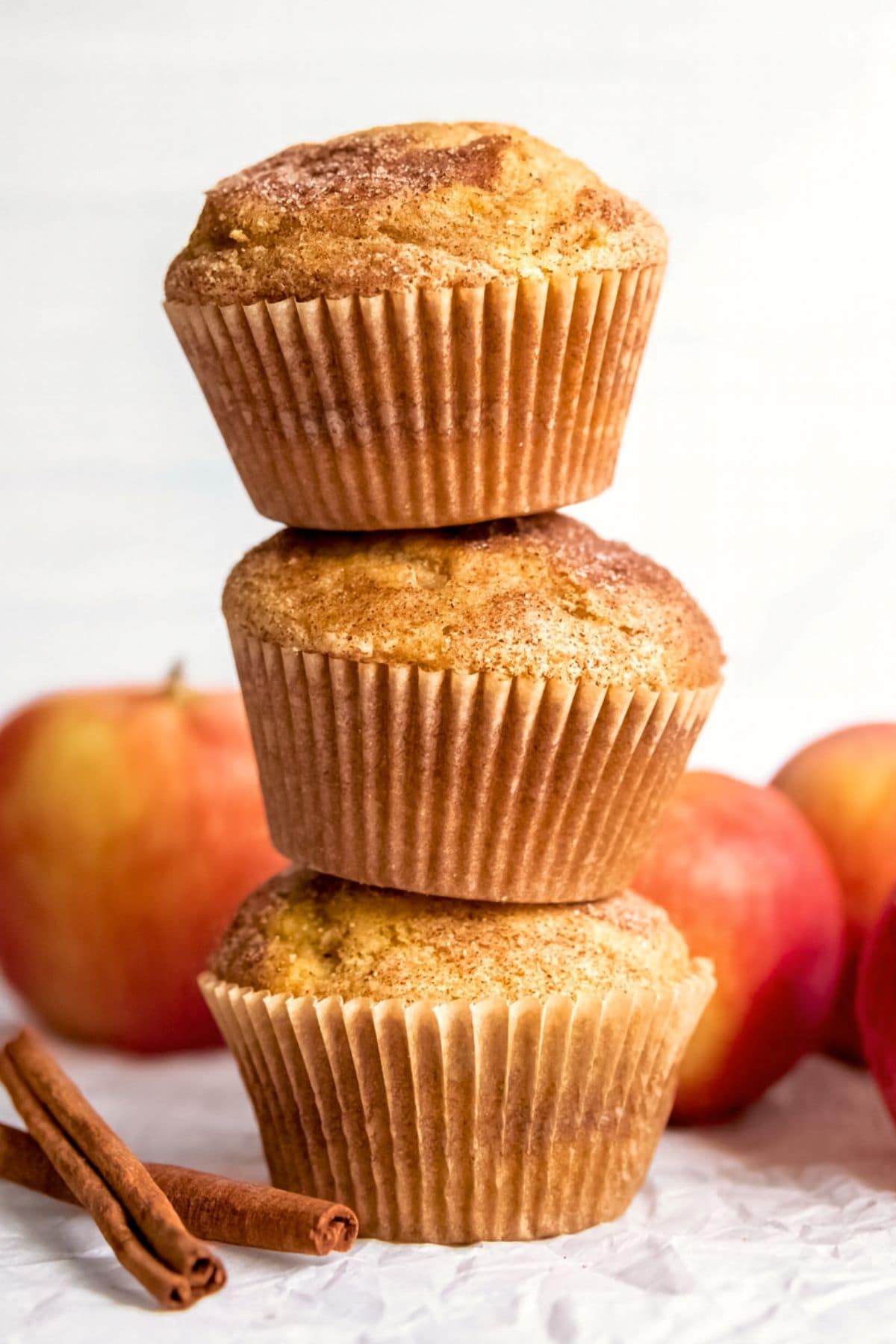 Stack of three apple cinnamon muffins next to cinnamon sticks and whole apples.