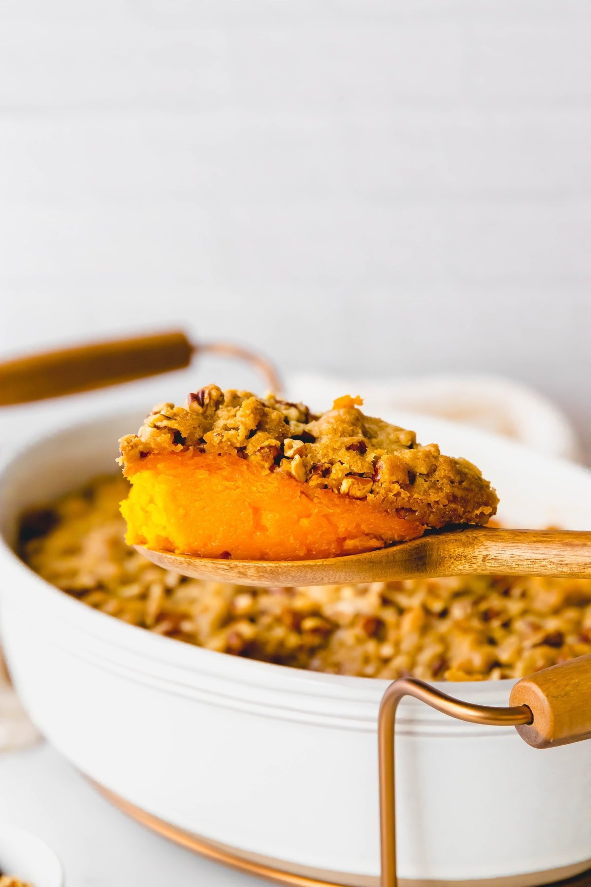 A wooden spoon holds a scoop of sweet potato casserole in front of the white casserole dish.
