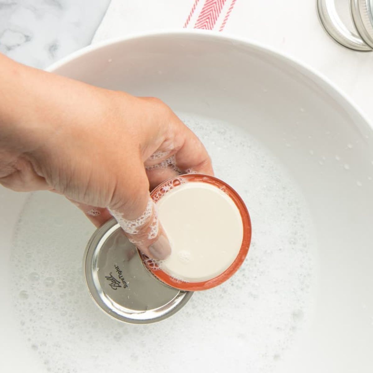 A hand washes flat jar lids in a bowl of soapy water.